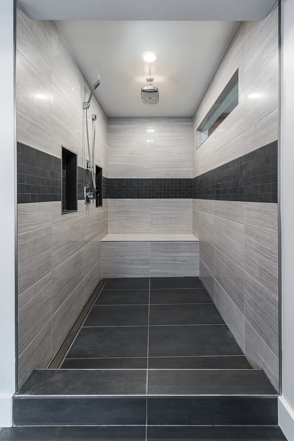 Walk-in shower finished in soft grey tile with a dark accent band and a built-in bench