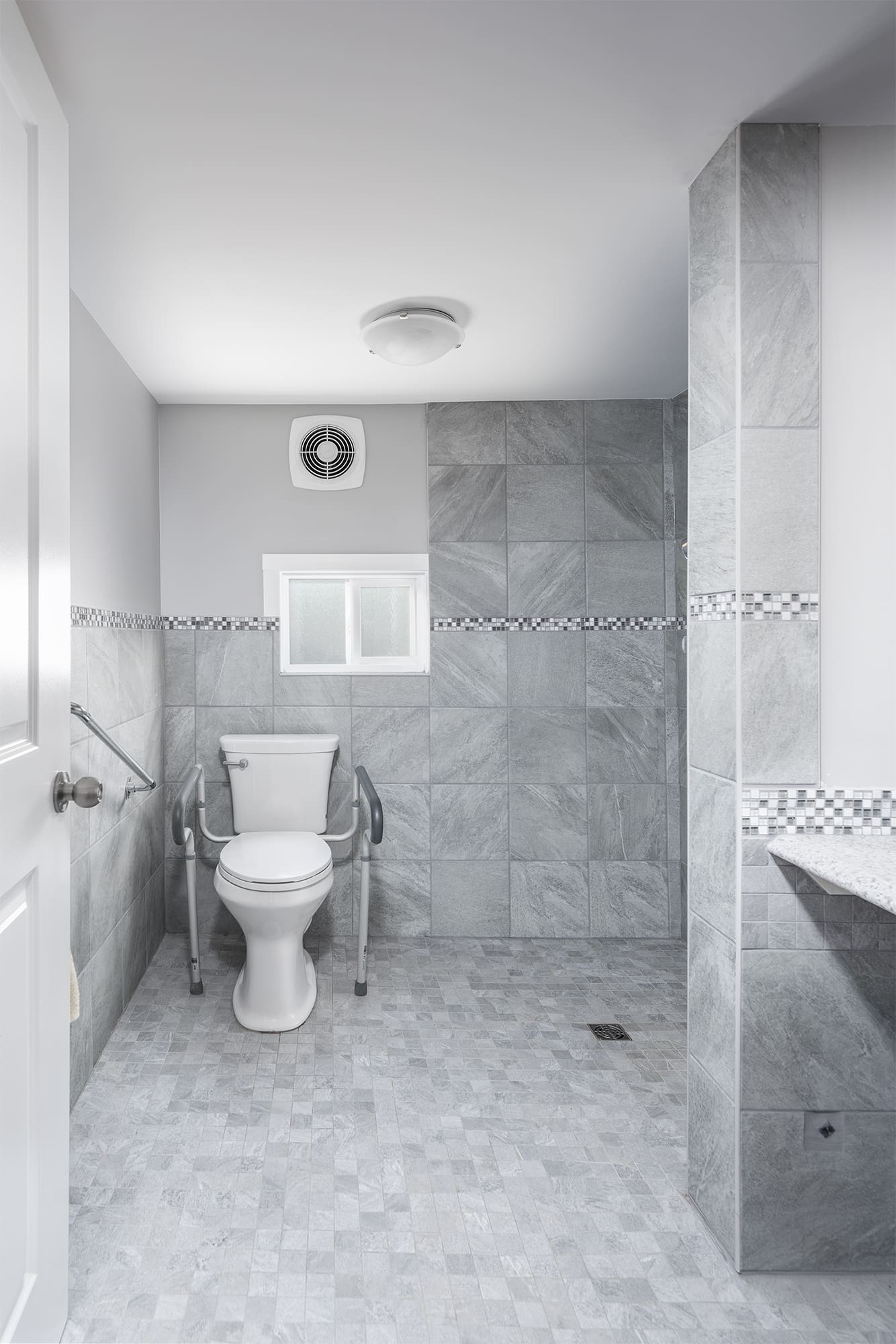 Accessible bathroom with comfort-height toilet, safety rails, curbless walk-in shower with built-in bench, and slip-resistant grey tile flooring