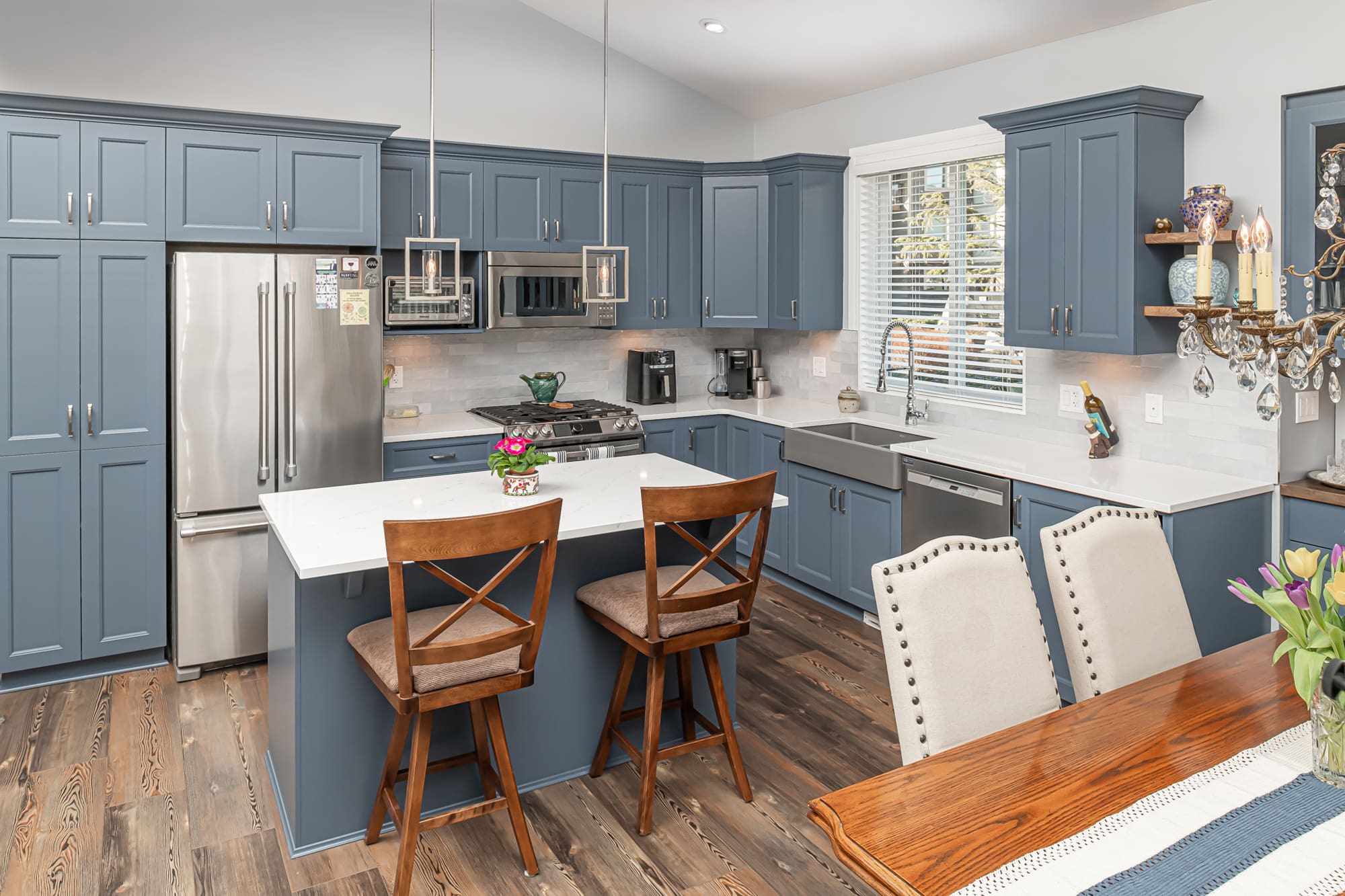 Elegant blue-grey kitchen renovation in Chilliwack featuring shaker cabinets, white quartz countertops, stainless steel appliances, hardwood flooring, and breakfast bar seating with crystal chandelier accent