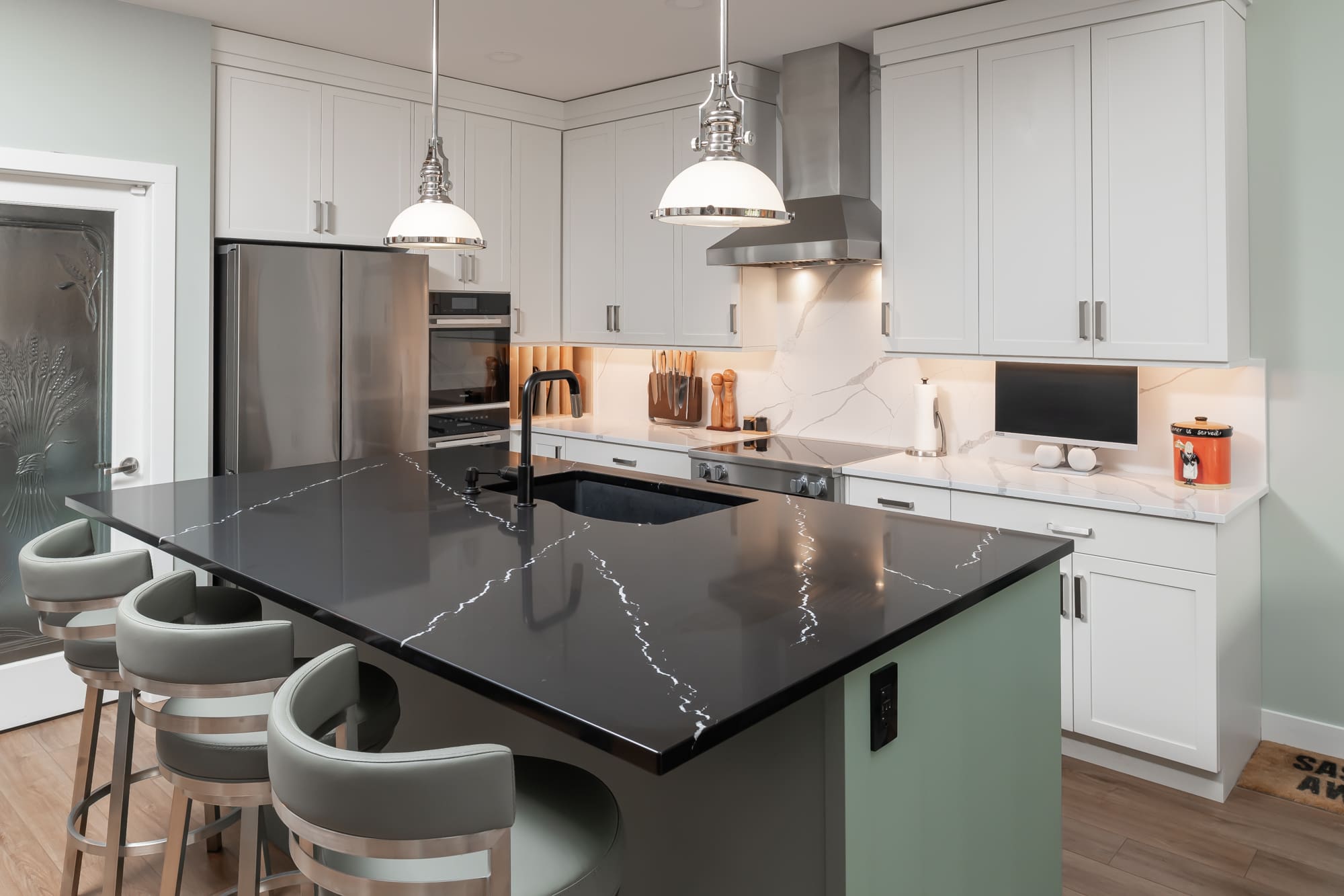 Modern Fraser Valley kitchen renovation featuring sage green island with black quartz countertop, white shaker cabinets, industrial pendant lights, stainless steel appliances, and marble backsplash designed for family connection and entertaining