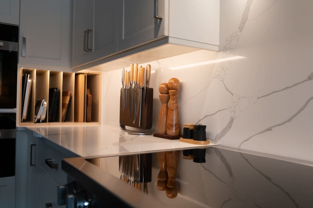 Under-cabinet lighting illuminating white quartz backsplash and knife block in a modern kitchen with vertical tray storage and natural wood accents