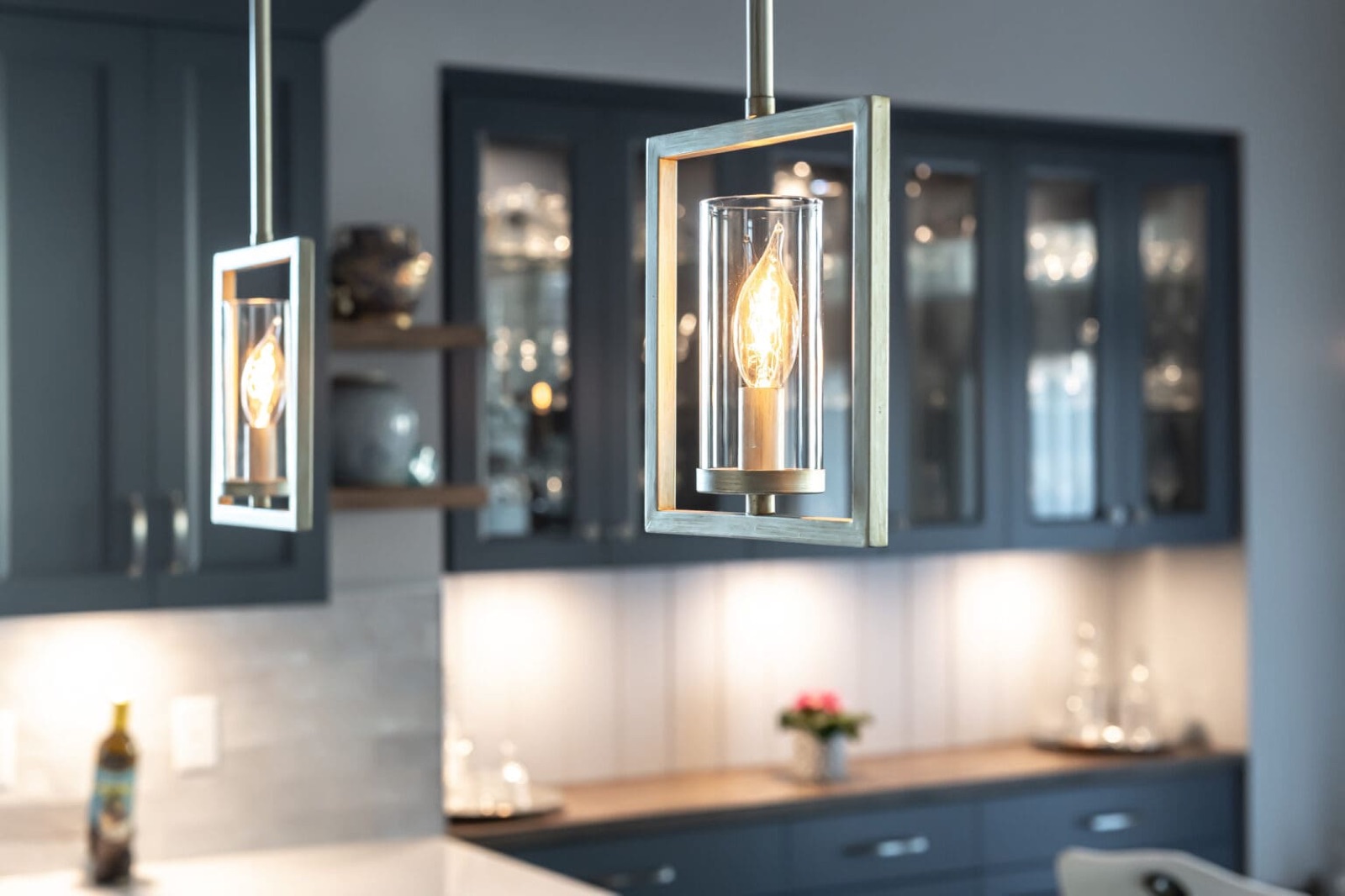 Modern lantern-style pendant lights with Edison bulbs above blue kitchen cabinetry with glass doors and under-cabinet lighting